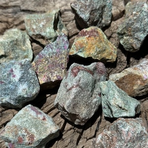 May include: A collection of rough, green, and gray stones with red and gold flecks. The stones are scattered on a wooden surface.