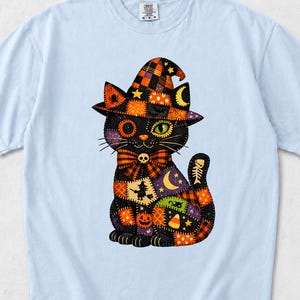 Puede incluir: Una camiseta azul claro con el gráfico de un gato negro que lleva un sombrero de bruja. El gato presenta un diseño de patchwork en tonos naranja, morado y negro, con motivos de calabazas, lunas, estrellas y una bruja sobre una escoba.