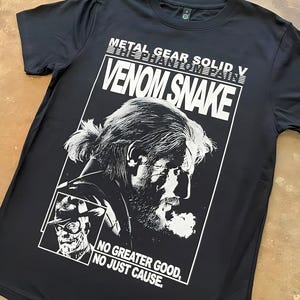 May include: Black t-shirt featuring a white graphic of "VENOM SNAKE" from Metal Gear Solid V. The design includes a portrait of a bearded man and the text "NO GREATER GOOD, NO JUST CAUSE."