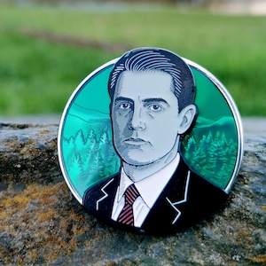 以下が含まれることがあります： スーツとネクタイ姿の男性の肖像画が描かれた丸いエナメルピン。肖像画は、森の風景を背景にしたティール色の背景に設定されています。ピンは銀色の縁取りがあり、石の表面に置かれています。