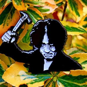 May include: Black and white enamel pin of a person holding a hammer. The person has dark hair and a serious expression. The pin is set against a background of yellow and green leaves.