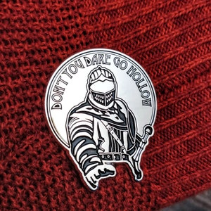 May include: A silver pin with a black and white illustration of a knight with the text "Don't You Dare Go Hollow" around the edge.