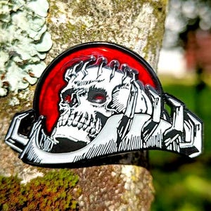 Może przedstawiać: Czarny, biały i czerwony pin emaliowany z motywem czaszki. Czaszka ma czerwone oczy i jest umieszczona na czerwonym tle. Pin jest przymocowany do pnia drzewa, z rozmytym zielonym tłem.