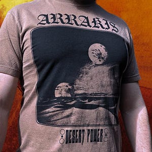 以下が含まれることがあります： ゴシック体の「ARRARIS」の文字がプリントされた茶色のTシャツ。黒い長方形には、二つの月と「DESERT POWER」の文字が描かれた砂漠の風景が描かれています。