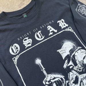 May include: Black long-sleeved t-shirt with a white graphic print of a knight holding a sword. The text "Knight of Astora Oscar" is printed in white on the shirt.
