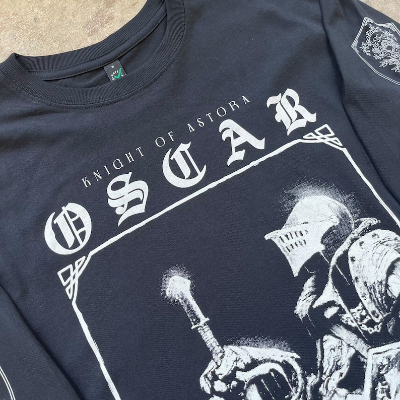 May include: Black long-sleeved t-shirt with a white graphic print of a knight holding a sword. The text "Knight of Astora Oscar" is printed in white on the shirt.