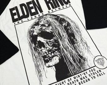 May include: Black and white graphic t-shirt with a black and white illustration of a skull with long hair. The text "Elden Ring The Lands Between" is printed on the shirt. The text "On a night of wintry fog the rune of death was stolen the demigods began to fall" is printed at the bottom of the shirt.