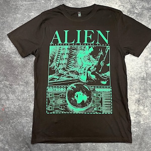 Könnte beinhalten: Schwarzes T-Shirt mit einem grünen Grafikdruck des Alien-Filmplakats. Die Grafik enthält den Titel "Alien", "Regie: Ridley Scott" und eine Szene aus dem Film.