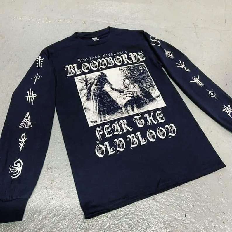 May include: Black long-sleeve shirt with white print. The print features the text "Hidetaka Miyazaki's Bloodborne" and "Fear the Old Blood". The sleeves have white graphic designs.