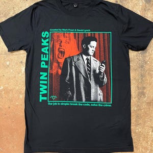 May include: Black t-shirt featuring a graphic from the TV show Twin Peaks. The design includes the show's title in green, a portrait of a man, and the text "Our job is simple: break the code, solve the crime."