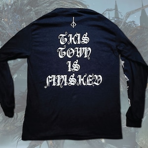 May include: Black long-sleeve shirt with white text that reads "THIS TOWN IS F*MSHED" in a gothic font. The shirt has a small symbol on the top center of the back.