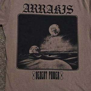 以下が含まれることがあります： 「ARRARIS」の文字が大きくスタイリッシュにプリントされた茶色のTシャツ。砂漠の風景と二つの月を描いた白黒のグラフィックが特徴。グラフィックの下には「DESERT POWER」の文字がプリントされています。