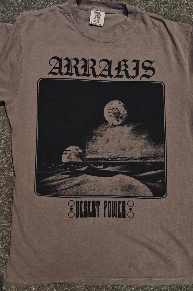 以下が含まれることがあります： 「ARRARIS」の文字が大きくスタイリッシュにプリントされた茶色のTシャツ。砂漠の風景と二つの月を描いた白黒のグラフィックが特徴。グラフィックの下には「DESERT POWER」の文字がプリントされています。
