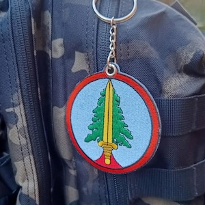 Pode incluir: Um porta-chaves redondo com fundo azul, apresentando uma árvore verde e uma espada amarela. O porta-chaves tem uma borda vermelha e é preso a uma corrente e um anel prateados. O fundo é uma mochila camuflada.