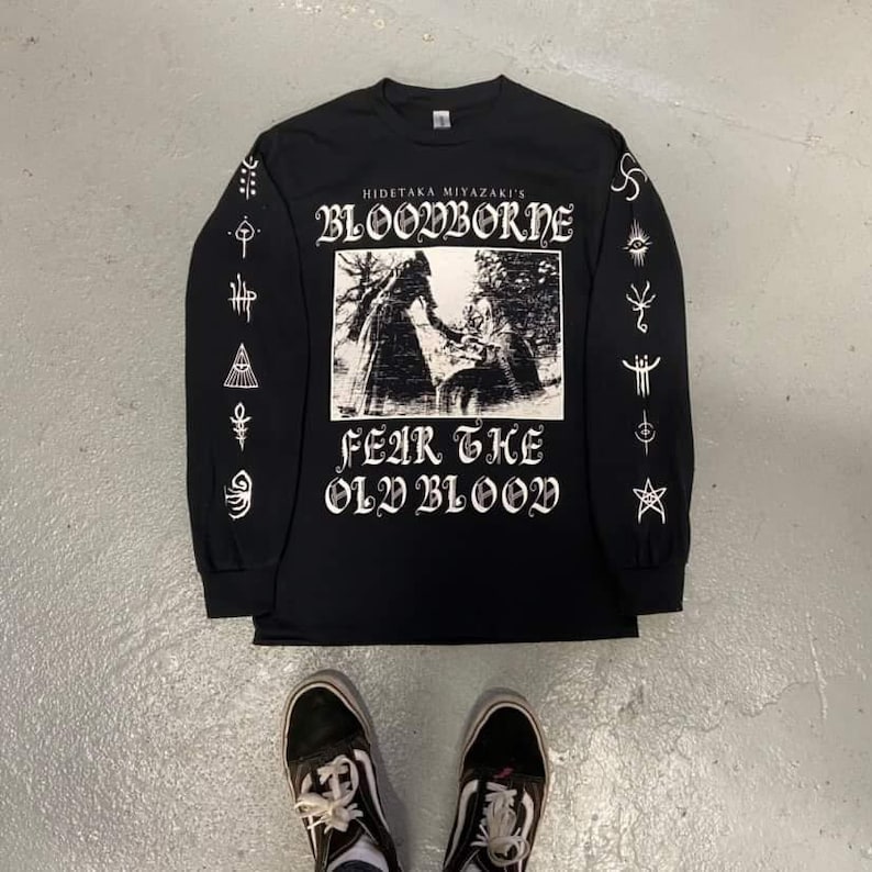May include: Black long-sleeve shirt with a white graphic print of a character from the video game Bloodborne. The text "Hidetaka Miyazaki's Bloodborne" and "Fear the Old Blood" are printed on the shirt.