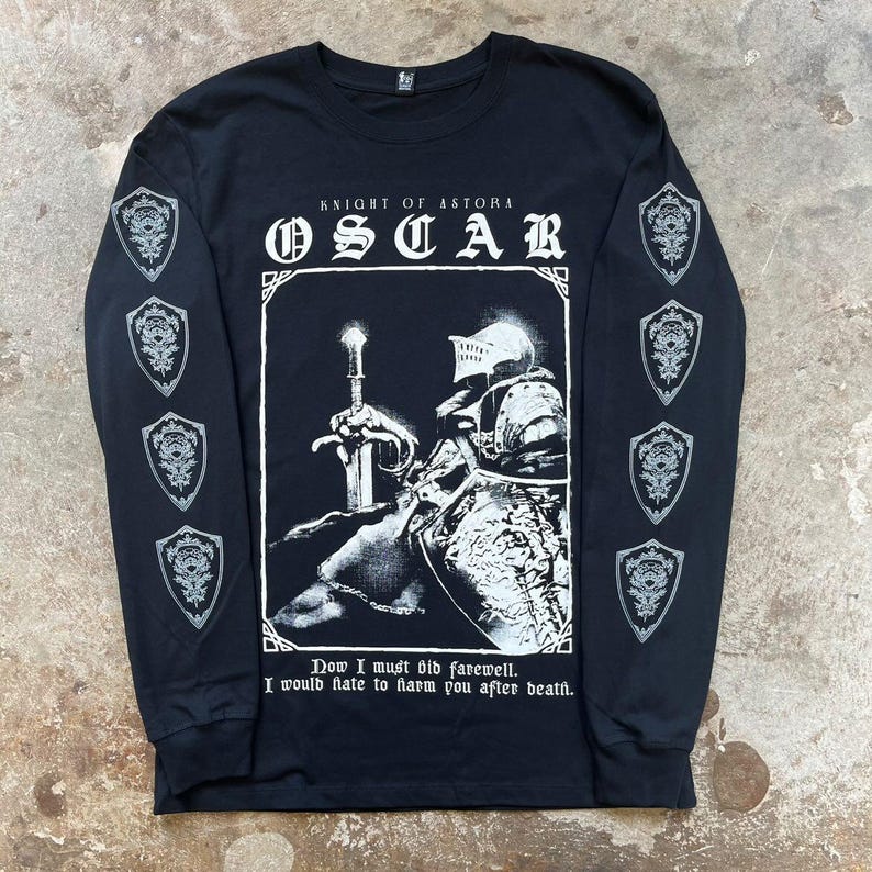 May include: Black long-sleeved t-shirt featuring a white graphic of a knight holding a sword. The graphic includes the text "Knight of Astora" and "Oscar". The text "Now I must bid farewell. I would hate to harm you after death." is printed below the graphic.