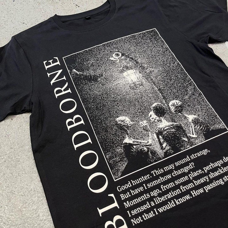 May include: Black t-shirt with a white graphic print of a scene from the video game Bloodborne. The graphic features a group of figures standing in front of a glowing light source. The text "Good hunter. This may sound strange, But have I somehow changed? Moments ago, from some place, perhaps de I sensed a liberation from heavy shackles Not that I would know. How passing st" is printed below the graphic.