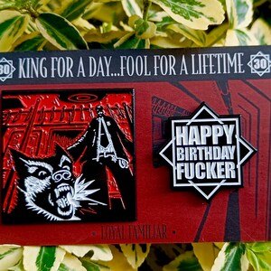 Può includere: Due spille smaltate su sfondo rosso. Una spilla presenta un'illustrazione in bianco e nero di un lupo e una figura con un lungo cappotto. L'altra spilla ha il testo "HAPPY BIRTHDAY FUCKER" in bianco. Il testo in alto recita "KING FOR A DAY...FOOL FOR A LIFETIME".