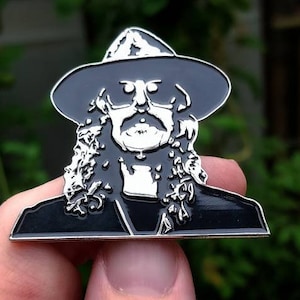 Puede incluir: Pin de esmalte en blanco y negro con el retrato de un hombre con el pelo largo y un sombrero. El pin es de color plateado y tiene un acabado brillante.