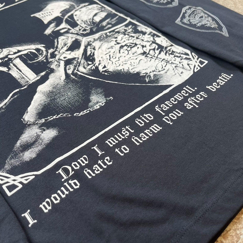 May include: A dark grey long-sleeved shirt with a white graphic print of a knight in armour. The text "Now I must bid farewell. I would hate to harm you after death." is printed on the shirt.