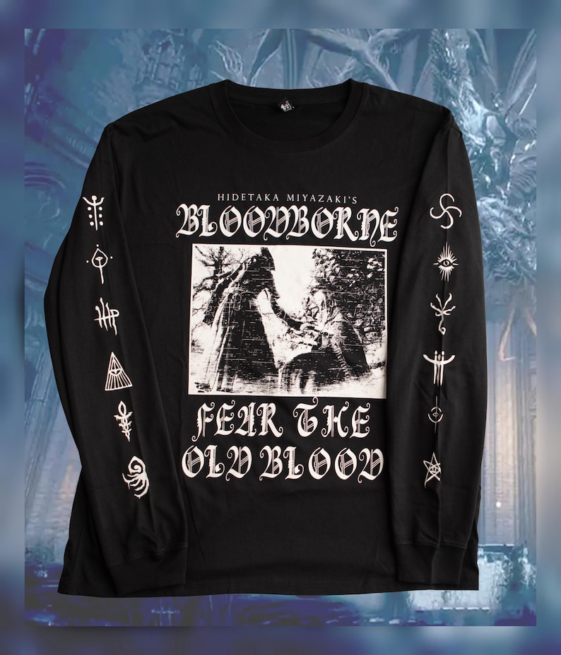 May include: Black long-sleeve t-shirt with a white graphic print of a scene from the video game Bloodborne. The text "Hidetaka Miyazaki's Bloodborne" and "Fear the Old Blood" are printed on the shirt. The sleeves feature white graphic prints of symbols.
