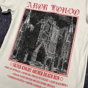Könnte beinhalten: Ein weißes T-Shirt mit einem Schwarzweißbild einer gotischen Kathedrale. Der Text "ANOR LONDO" ist in Rot oben auf dem Bild gedruckt. Unter dem Bild ist der Text "OSILVER KNIGHT ARCHER DEATH RUNO" in Rot gedruckt. Darunter ist eine Liste von Charakteren aus dem Videospiel Dark Souls in Schwarz gedruckt.