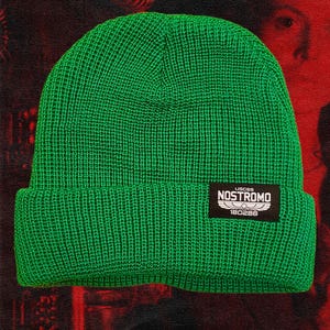May include: Bright green knit beanie with a folded brim. A black rectangular patch on the brim displays the text "USCSS NOSTROMO" and the number "180286" in white. The background is a dark red and black.