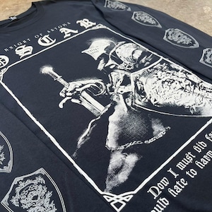 May include: Black long-sleeved t-shirt featuring a white graphic print of a knight in armour holding a sword. The text "Knight of Astora" and "OSCAR" are printed on the shirt. The text "Dow I must bid farewell to hate to harm" is printed at the bottom of the graphic.