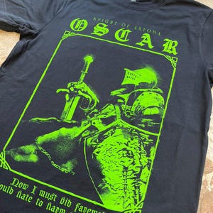 Könnte beinhalten: Schwarzes T-Shirt mit einem grünen Grafikdruck eines Ritters in Rüstung, der ein Schwert hält. Der Text "Knight of Astora" und "Oscar" ist über dem Ritter gedruckt. Der Text "Now I must bid farewell. Would hate to harm you after death." ist unter dem Ritter gedruckt.