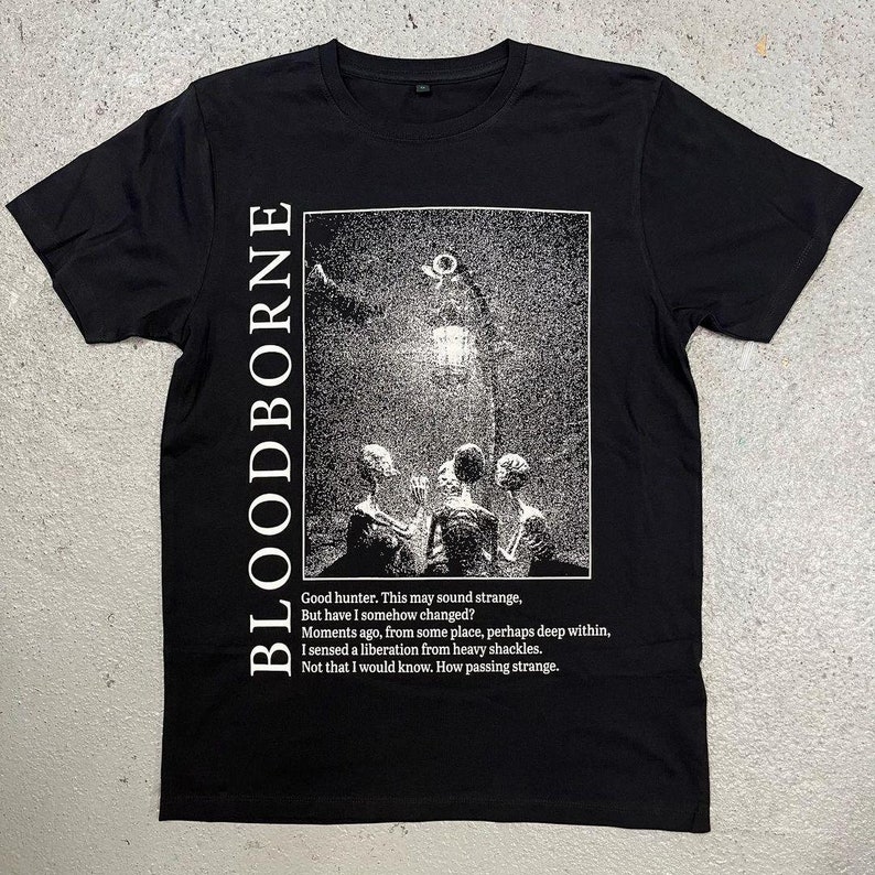 May include: Black t-shirt with a white graphic print of a scene from the video game Bloodborne. The graphic features a group of figures standing in front of a large, glowing lantern. The text "Good hunter. This may sound strange, But have I somehow changed? Moments ago, from some place, perhaps deep within, I sensed a liberation from heavy shackles. Not that I would know. How passing strange." is printed below the graphic.