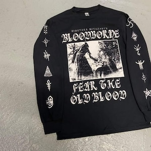 May include: Black long-sleeve t-shirt with a white graphic print of a scene from the video game Bloodborne. The text "Hidetaka Miyazaki's Bloodborne" and "Fear the Old Blood" are printed on the shirt. The sleeves feature various white symbols.