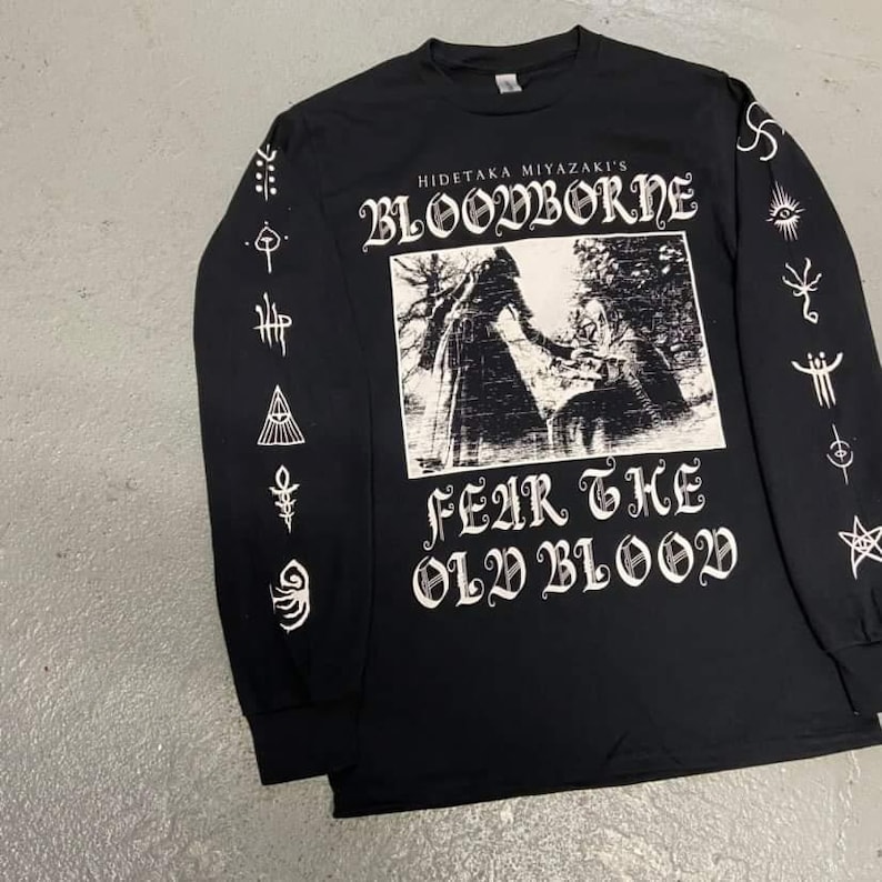 May include: Black long-sleeve t-shirt with a white graphic print of a scene from the video game Bloodborne. The text "Hidetaka Miyazaki's Bloodborne" and "Fear the Old Blood" are printed on the shirt. The sleeves feature various white symbols.