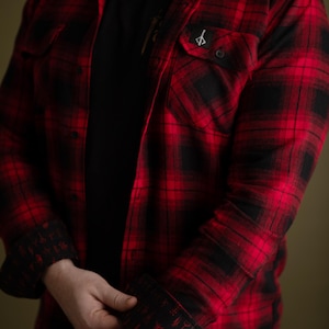 Op de afbeelding: Een rood en zwart geruit flanellen overhemd met een zwarte voering met een herhalend patroon van symbolen. Het overhemd heeft een enkele knoopsluiting op de linkerborst met een klein geborduurd symbool.