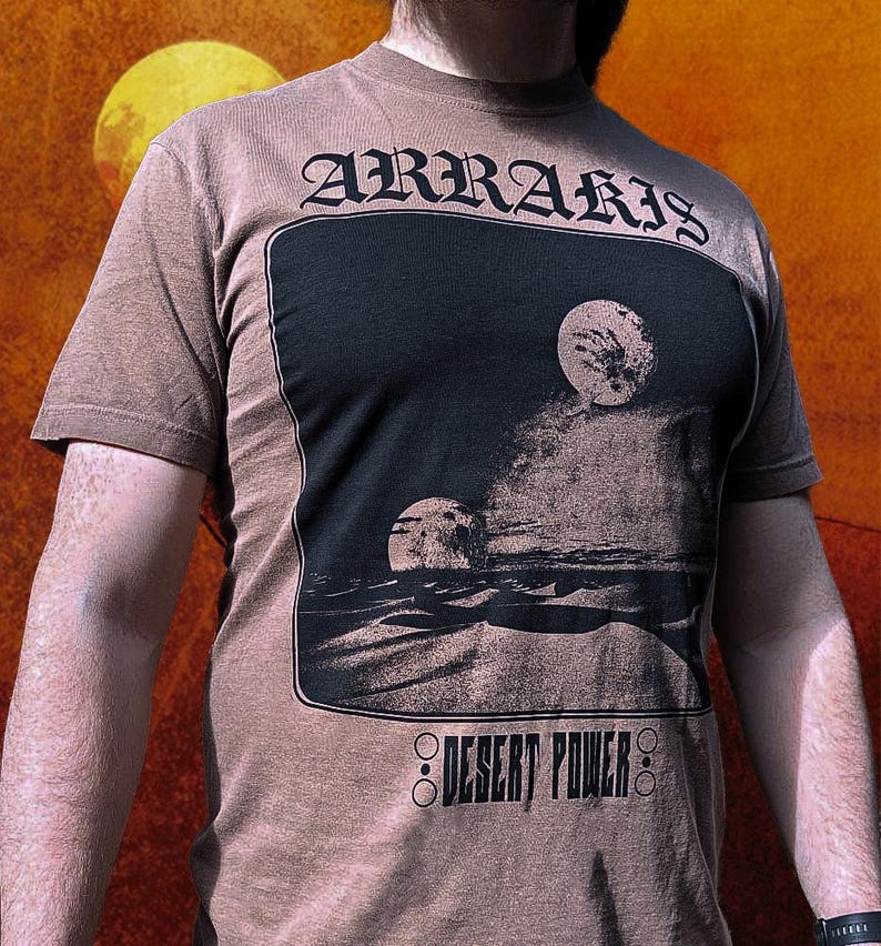 以下が含まれることがあります： ゴシック体の「ARRARIS」の文字がプリントされた茶色のTシャツ。黒い長方形には、二つの月と「DESERT POWER」の文字が描かれた砂漠の風景が描かれています。