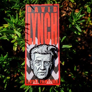 Puede incluir: Marcapáginas rectangular con un retrato de David Lynch y el texto "DAVID LYNCH" en letras rojas. El texto "LOYAL FAMILIAR" está en la parte inferior. El fondo es una planta verde.