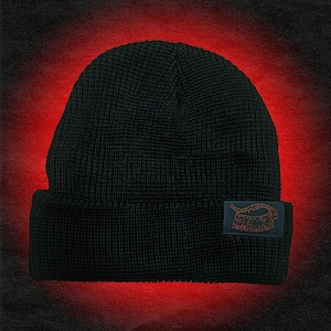 Op de afbeelding: Een zwarte gebreide beanie met een omgeslagen rand. Een klein rechthoekig label met een oranje ontwerp is aan de rand bevestigd. De achtergrond is een rood-zwarte gradiënt.