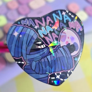May include: A heart-shaped holographic sticker featuring a blue and black mummy holding a pink and white ghost. The sticker has the text "NANANA" repeated around the edge.