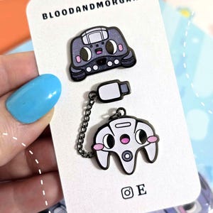 May include: Enamel pins featuring a cartoon-style video game controller and console. The controller is white with pink accents, and the console is gray. The pins are connected by a chain and are on a white card with the text "BLOODANDMORGANS".