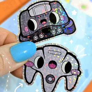 May include: Two holographic stickers featuring cartoon-style game consoles. The top sticker resembles a retro boombox, while the bottom one is a game controller. Both have black eyes, pink cheeks, and a silver glitter effect.