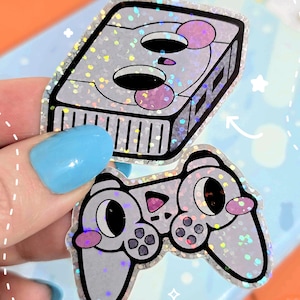 May include: Two stickers: a silver glittery video game console and a matching controller. The console has a rectangular shape with black details and pink accents. The controller has a cartoonish design with large eyes and buttons.
