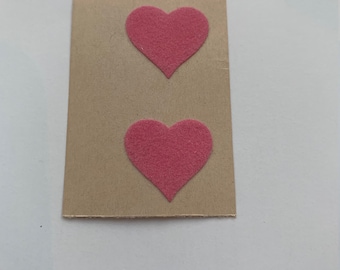May include: A rectangular piece of tan cardboard featuring two pink felt heart-shaped cutouts attached to the surface. The hearts are positioned vertically, one above the other, centered on the flat, light brown rectangular card base.