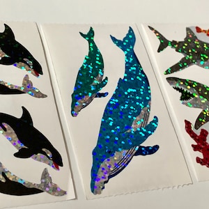 Könnte beinhalten: Drei Blätter mit Aufklebern, die verschiedene Meerestiere zeigen. Das erste Blatt zeigt zwei Orcas, das zweite Blatt zeigt zwei Wale und das dritte Blatt zeigt zwei Haie, ein Korallenriff und einen kleinen Fisch. Alle Aufkleber sind schillernd.