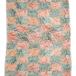 May include: A pink and green checkered rug with a fringe border. The rug is made of a soft, fluffy material.