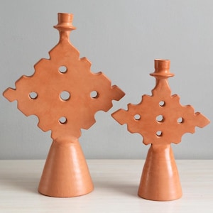 May include: Two terracotta candle holders with a smooth finish. Each piece features a wide, conical base supporting a flat, diamond-shaped top with jagged edges and five circular cutouts. A small cylindrical candle cup sits at the very top.