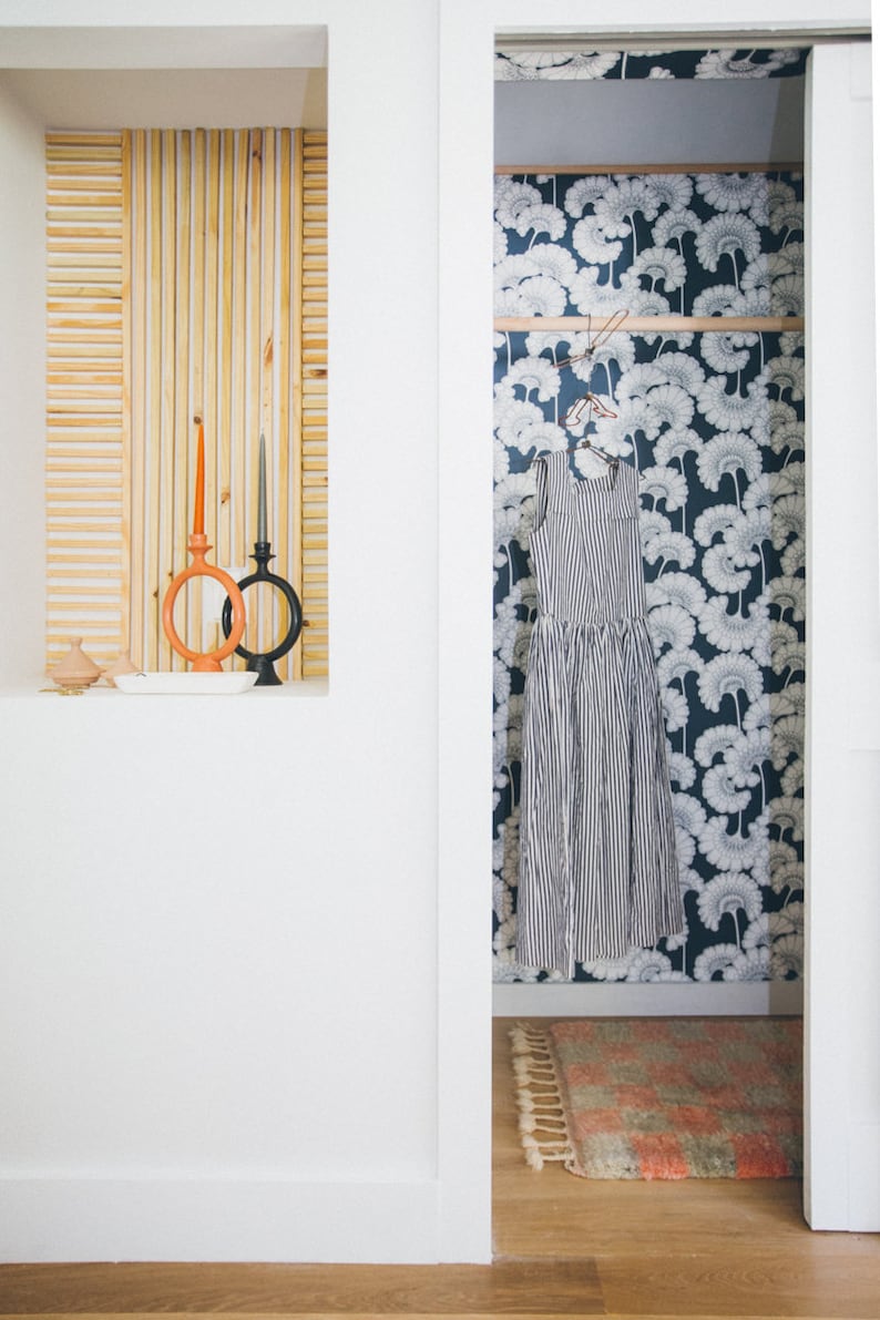 May include: A white closet door is open, revealing a closet with a blue and white floral wallpaper. A striped dress hangs on a clothes hanger inside the closet. A small rug with pink and white stripes is on the floor of the closet.