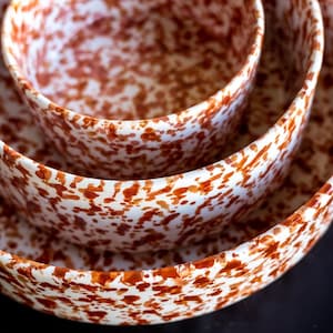 May include: A stack of three ceramic bowls with a speckled pattern. The bowls are white with reddish-brown spots. The bowls are nested inside each other, with the largest bowl at the bottom and the smallest at the top. The bowls are on a dark surface.