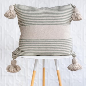 May include: A square throw pillow featuring a woven pattern of sage green and off-white horizontal stripes. The pillow has a wide solid off-white band across the center and is finished with four large, thick off-white tassels at each corner.
