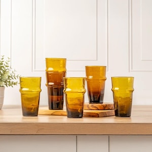 May include: A set of five amber-colored glass tumblers with a distinct horizontal ridge near the top rim. The glasses feature a translucent, warm brown hue and a cylindrical shape with a slightly tapered base, arranged in a group on a flat surface.