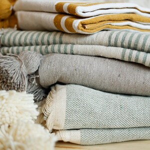 May include: A stack of folded towels in various colors and patterns, including white, gray, green, and yellow. The towels are made of a soft, textured fabric and have a fringed edge.