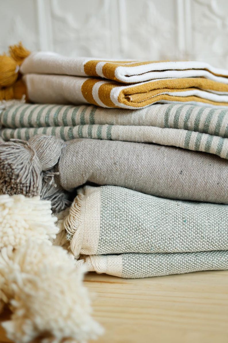 May include: A stack of folded towels in various colors and patterns, including white, gray, green, and yellow. The towels are made of a soft, textured fabric and have a fringed edge.
